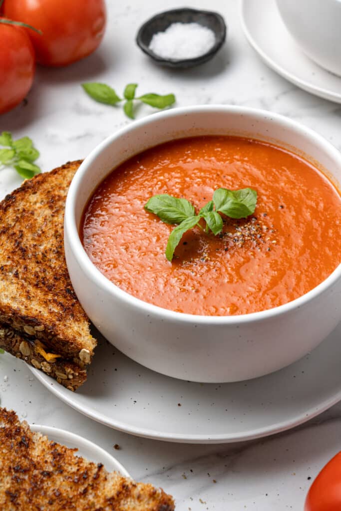 close up side view tomato soup in white bowl over plate with grilled cheese sandwich