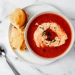 Overhead view of a bowl of tomato soup on top of a white plate with a sandwich on it beside a spoon.