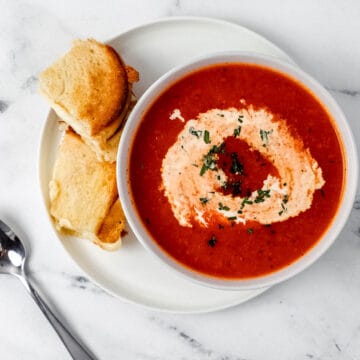 Overhead view of a bowl of tomato soup on top of a white plate with a sandwich on it beside a spoon.