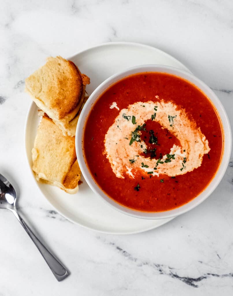 Overhead view of a bowl of tomato soup on top of a white plate with a sandwich on it beside a spoon.