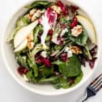 Over head view of salad in white bowl beside a fork on white surface.