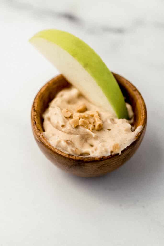 snickerdoodle dip in small wooden bowl with green apple slice and topped with toffee bits