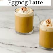 Close up side view of two finished eggnog lattes in glass mugs on pin with text overlay.