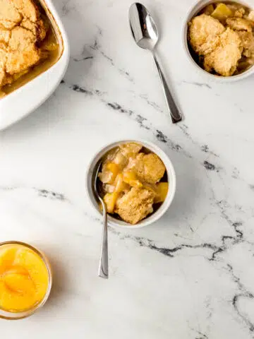 overhead view peach cobbler in small white bowls and spoons on marble surface