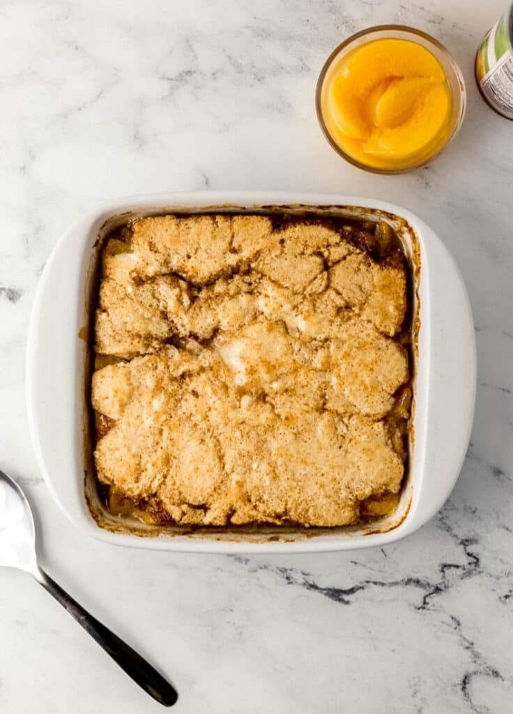 baked peach cobbler in square white baking dish with canned peaches and spoon