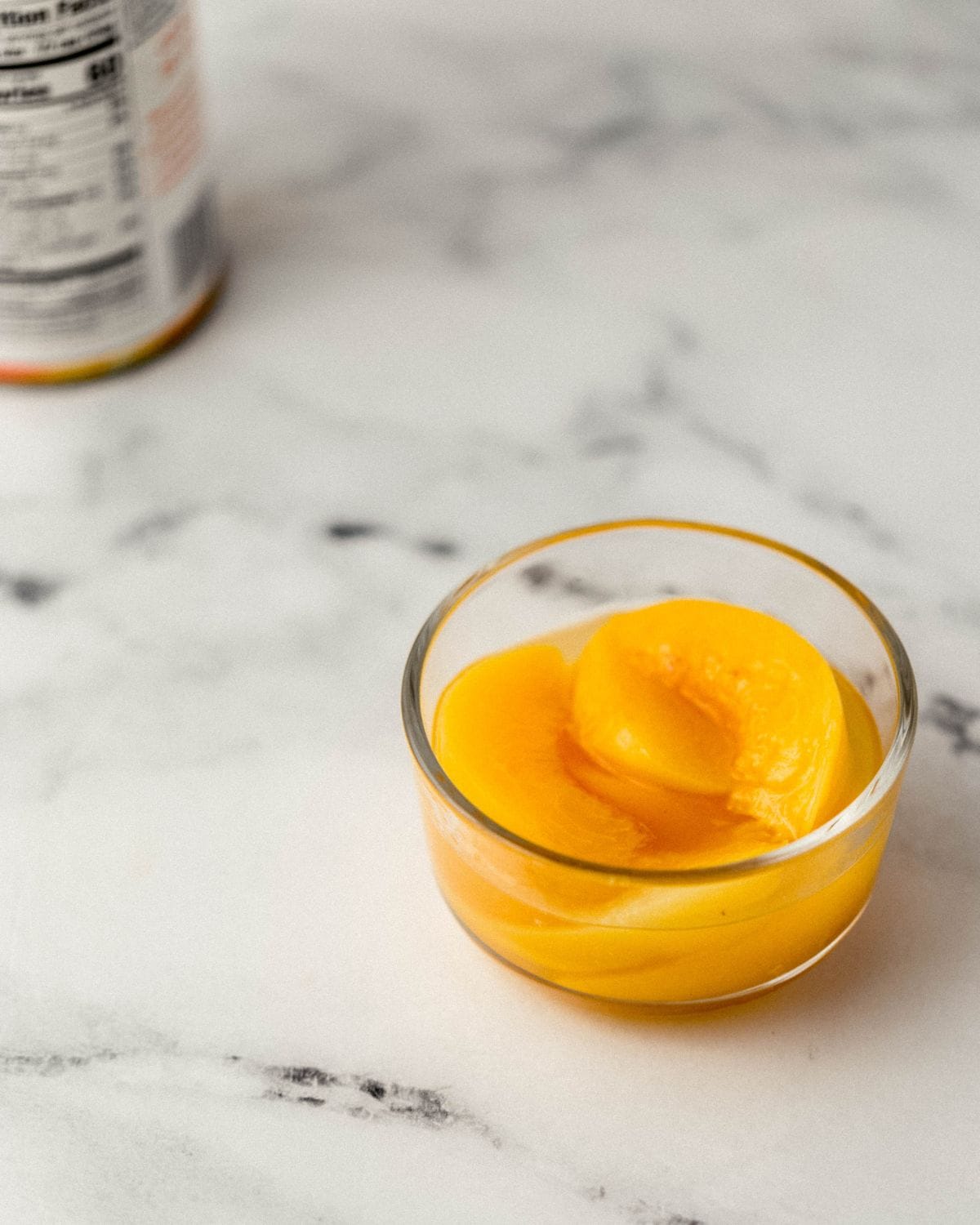 Close up front view of sliced canned peaches in a glass bowl with the can in the background. 
