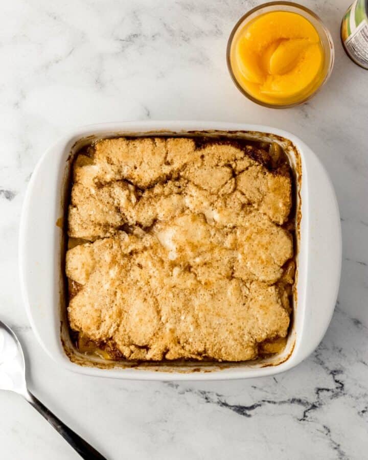Overhead view of baked peach cobbler in white baking dish by glass bowl of sliced canned peaches. 
