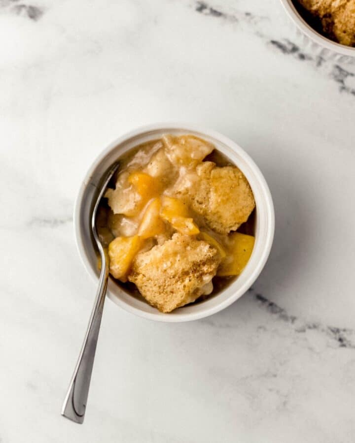 Close up overhead view of single serving of peach cobbler in white bowl with spoon in it.