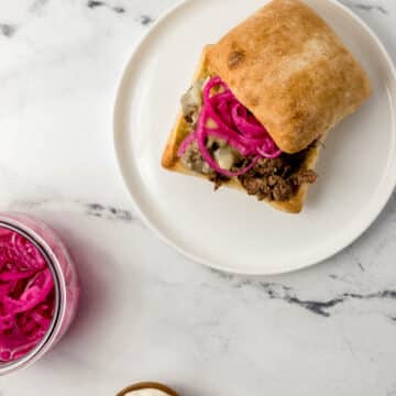 steak sandwich on white plate beside sauce and onions