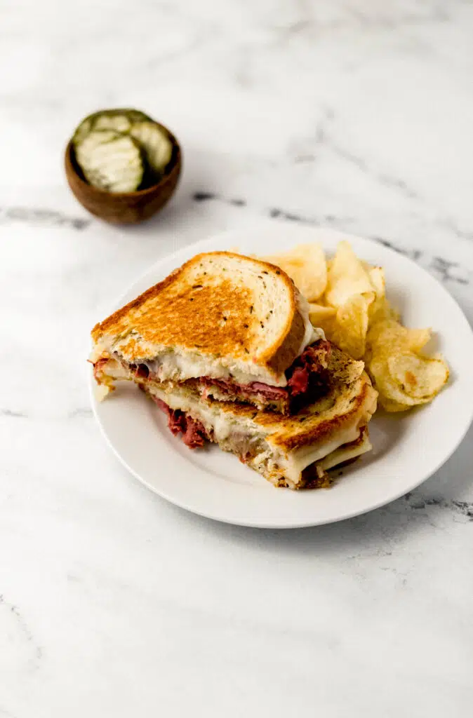 grilled cheese sandwich cut in half on white plate with chip and a small wooden bowl of pickles 