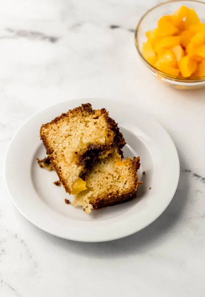 slice of peach bread on white plate with small glass bowl of diced peaches in the background
