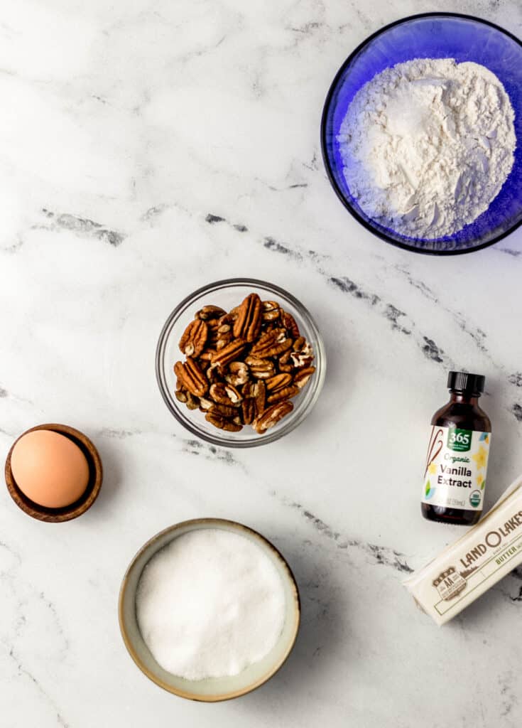 ingredients needed to make pecan sandies in separate containers on marble surface 