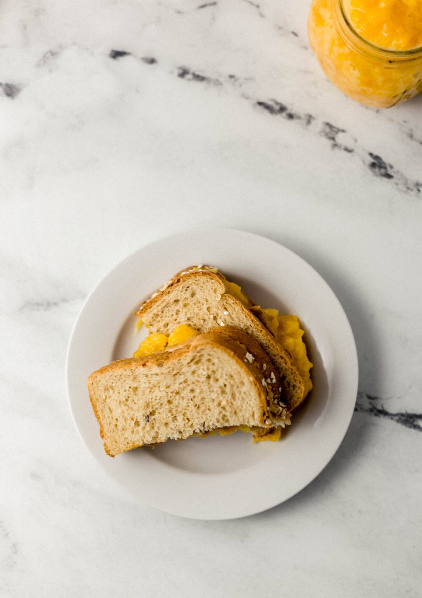 overhead view of sandwich cut in half on small white plate with jar of jam in the background