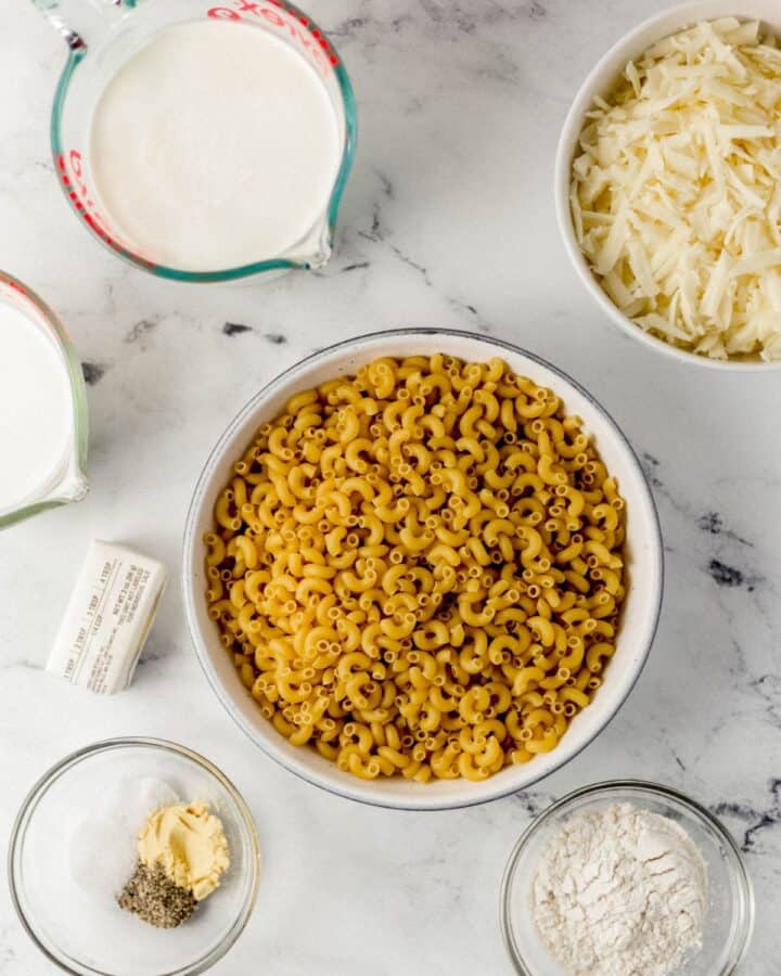 Ingredients needed to make mac and cheese in separate bowls on marble surface. 