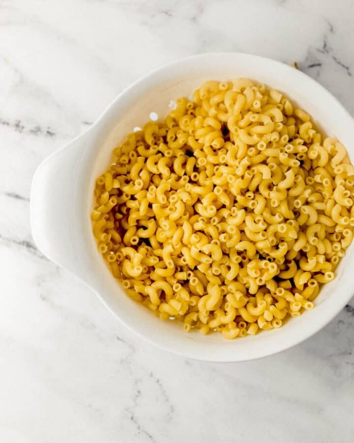 Cooked and drains elbow pasta in strainer on marble surface. 