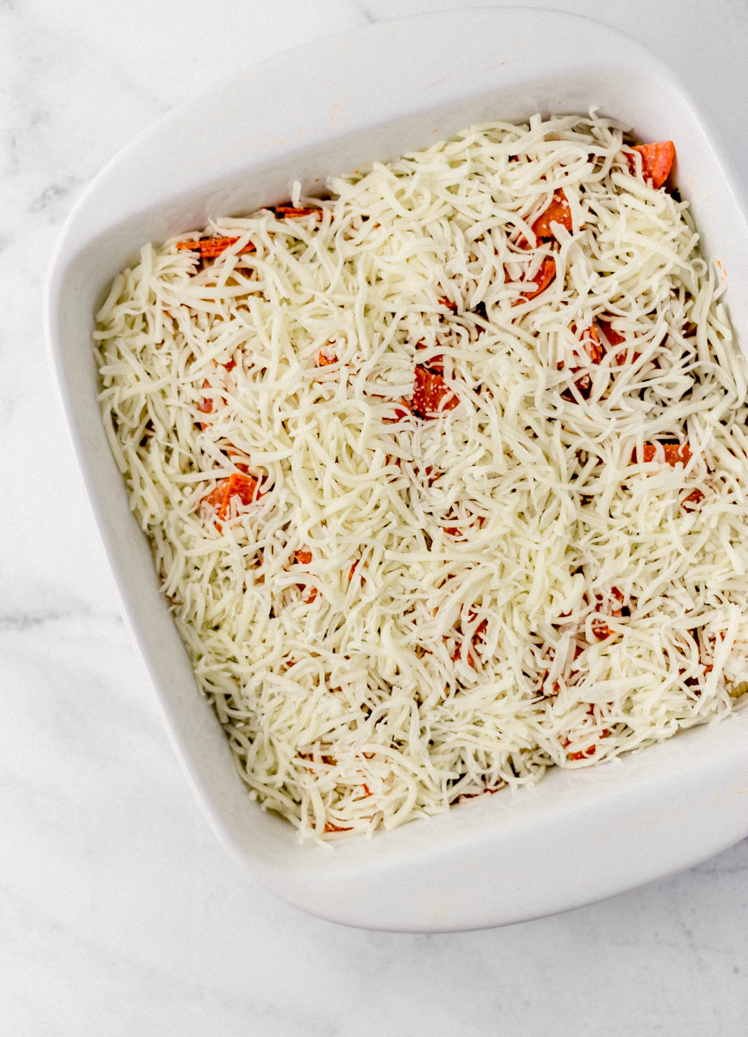 shredded cheese spread on top of pasta in white baking dish 