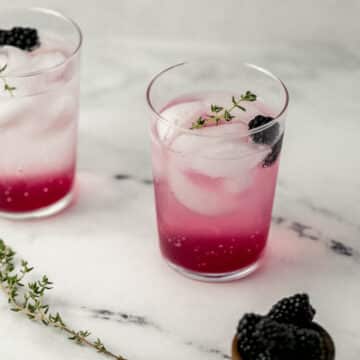 close up side view of drink in glass with ice, blackberry, and thyme