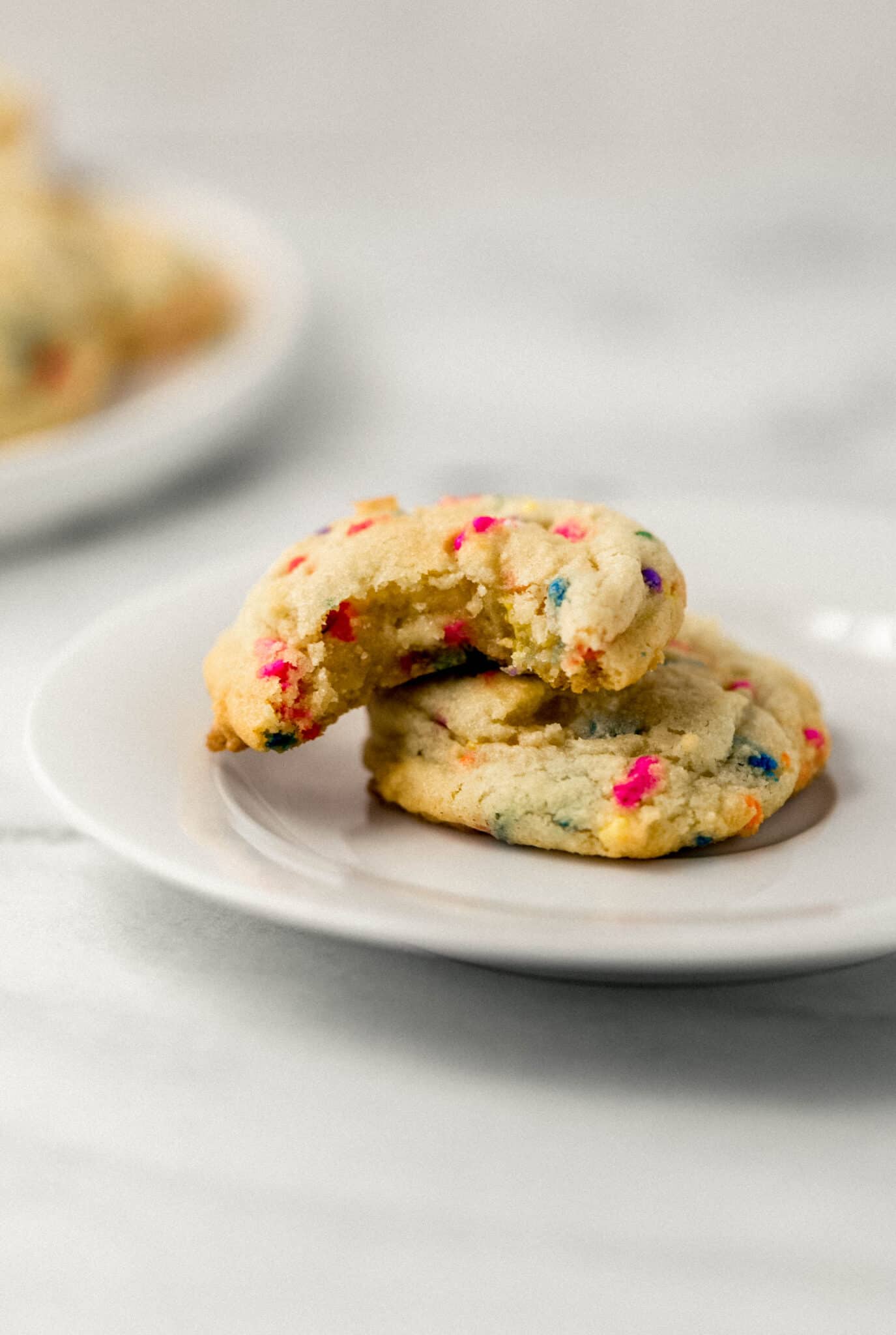 close up side view of two sugar cookies on white plate, one with bite in it. 