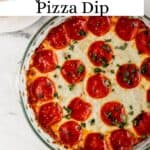 Overhead view of baked pizza dip in glass dish by plate of bread on pin with text overlay.