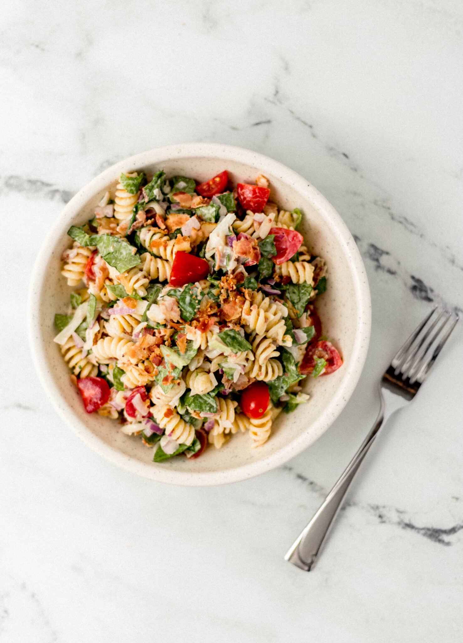 pasta salad in white bowl beside a fork on marble surface