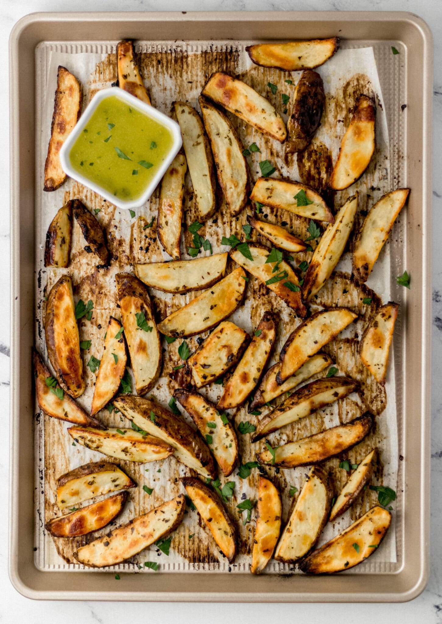 overhead view of cooked Greek potatoes on parchment lined baking sheet with square white container of sauce. 