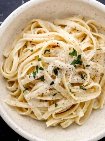 Overhead view of white bowl with pasta in it topped with parsley and black pepper on black tile surface.