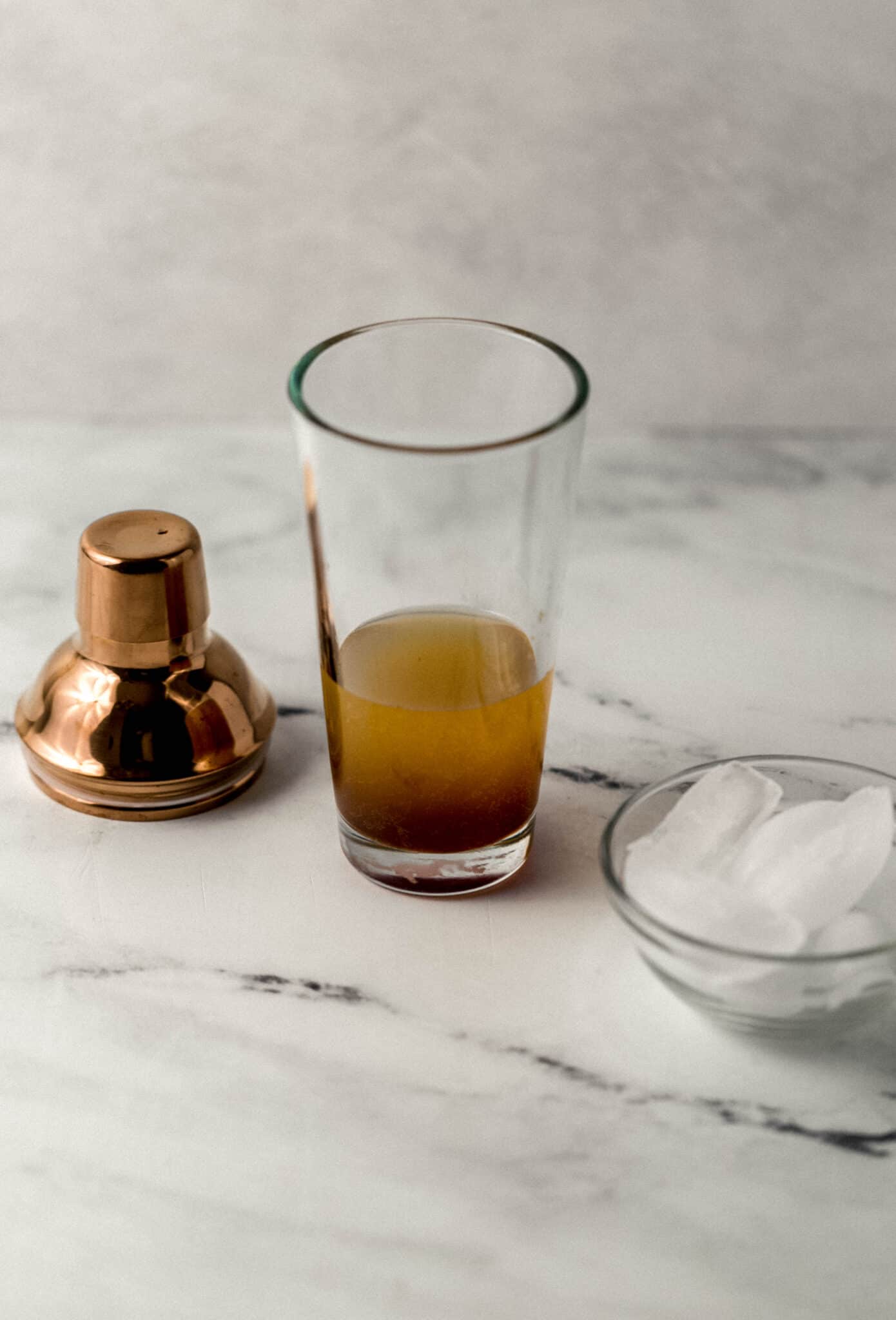 shaker glass with margarita ingredients in it beside shaker top and small glass bowl of ice