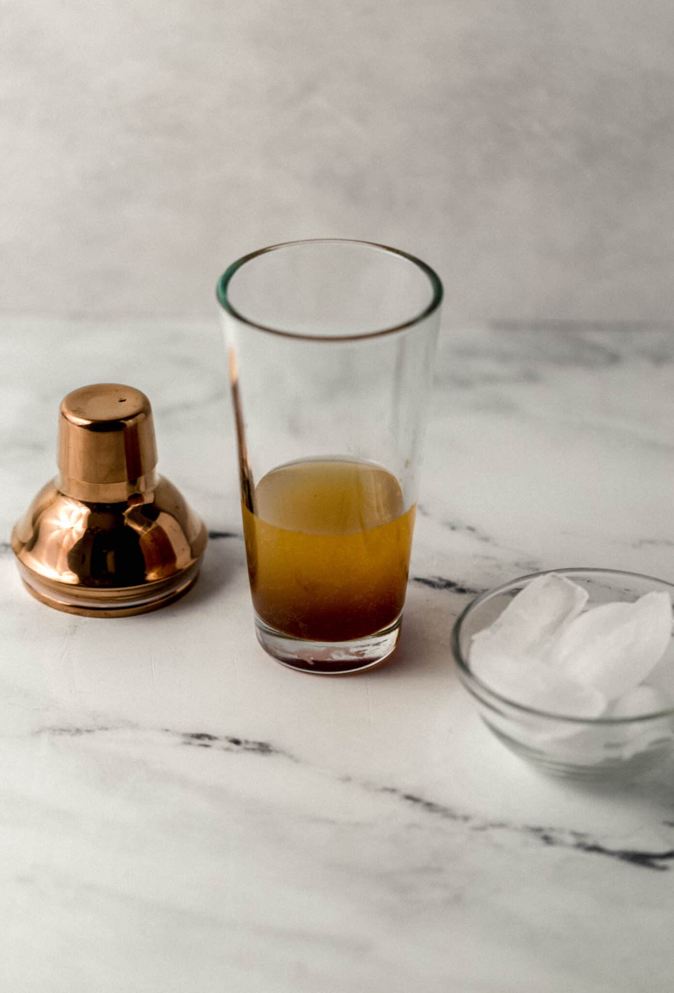 shaker glass with margarita ingredients in it beside shaker top and small glass bowl of ice 