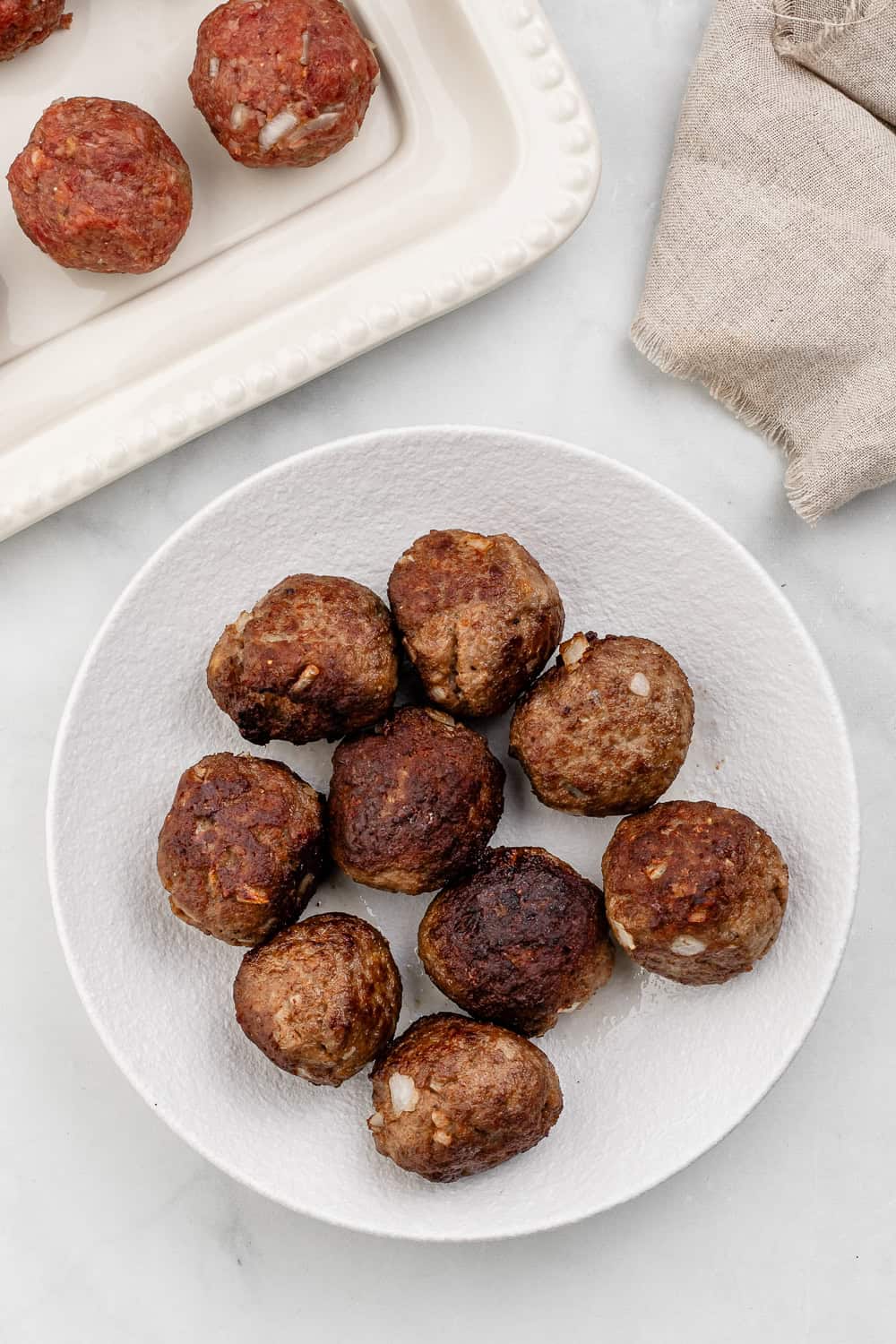 Overhead view of finished meatballs on a large white plate with uncooked meatballs in the background.