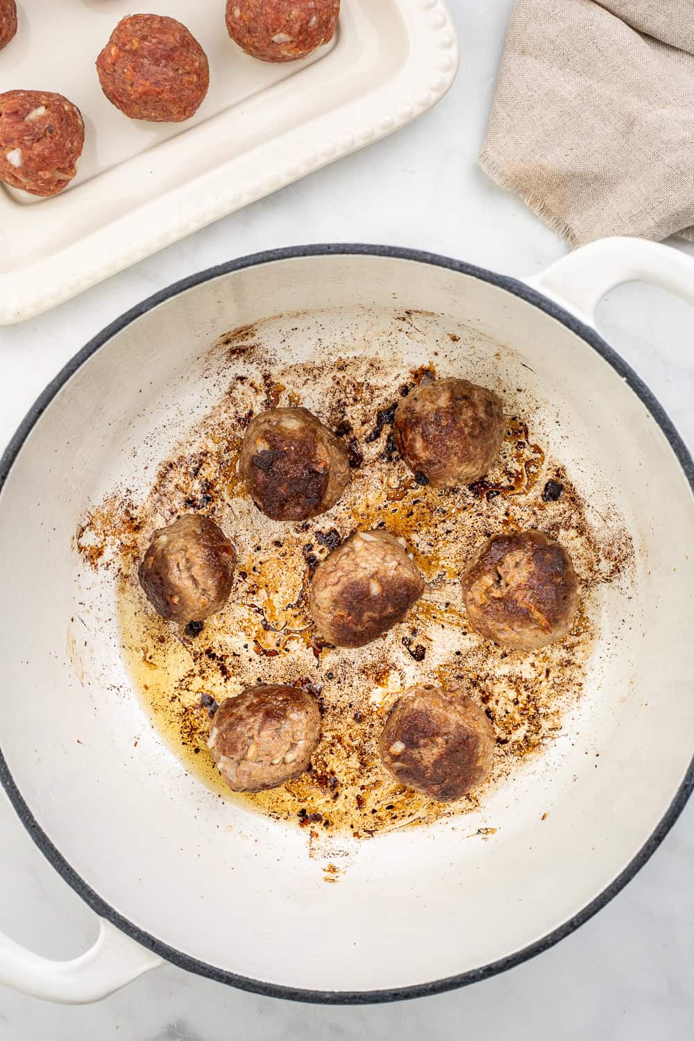Meatballs cooking in large white dutch oven with other meatballs in the background.