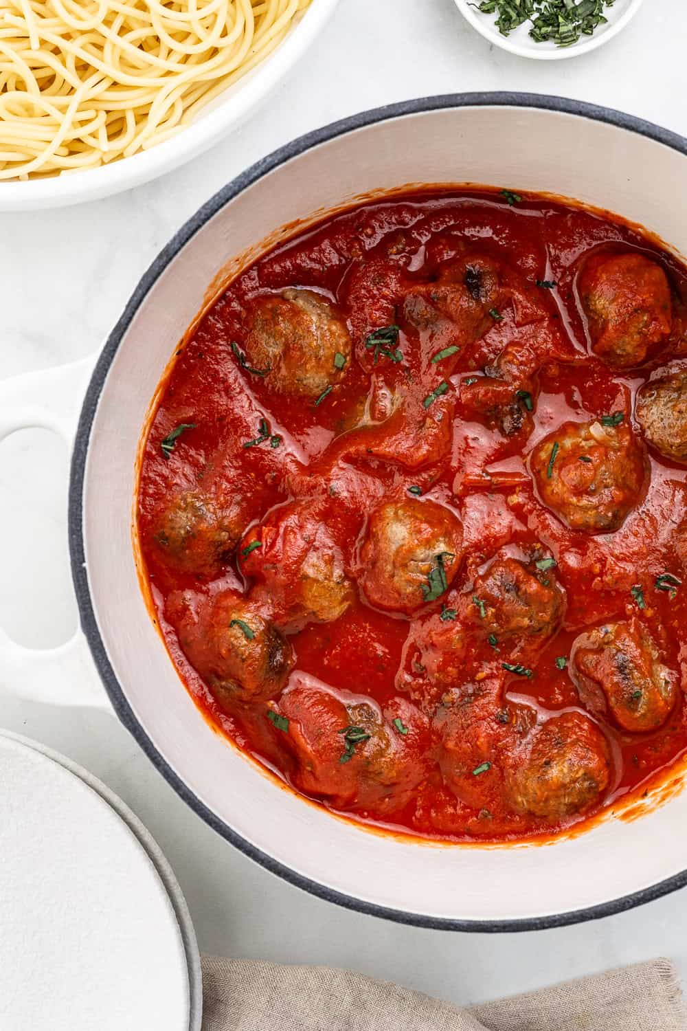 Meatballs and sauce in large white dutch oven by bowl of spaghetti.