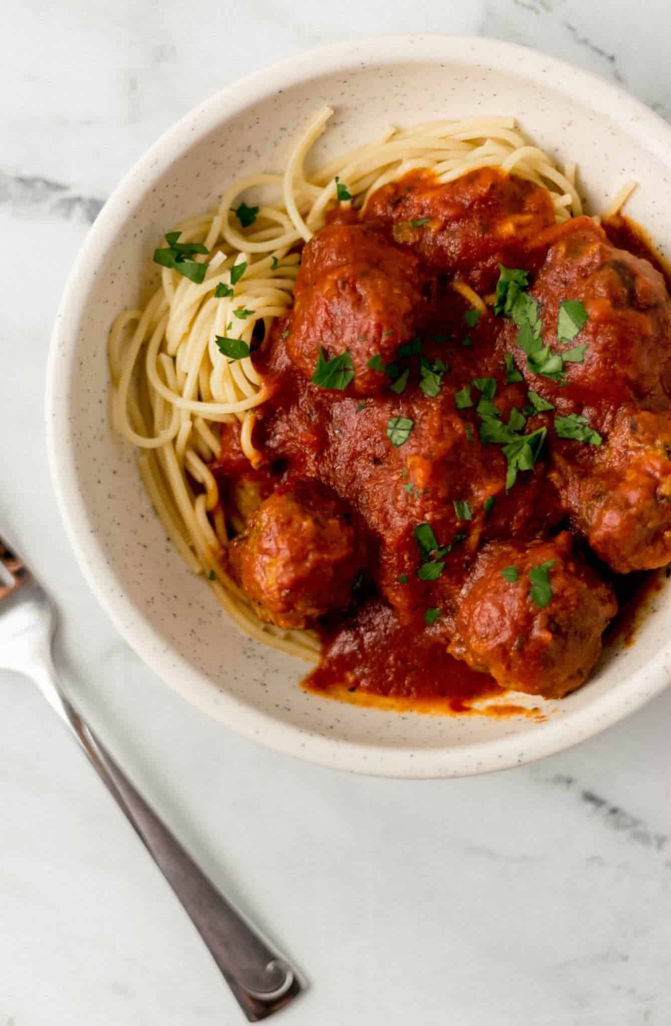 overhead view of white bowl of spaghetti and meatballs topped with parsley beside a fork