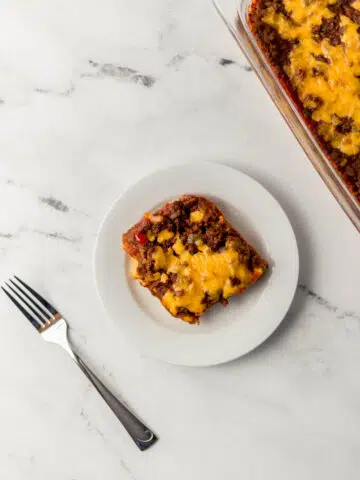 overhead view of sloppy joe cornbread casserole serving on a small white plate beside a fork and the rest of the casserole in a glass dish