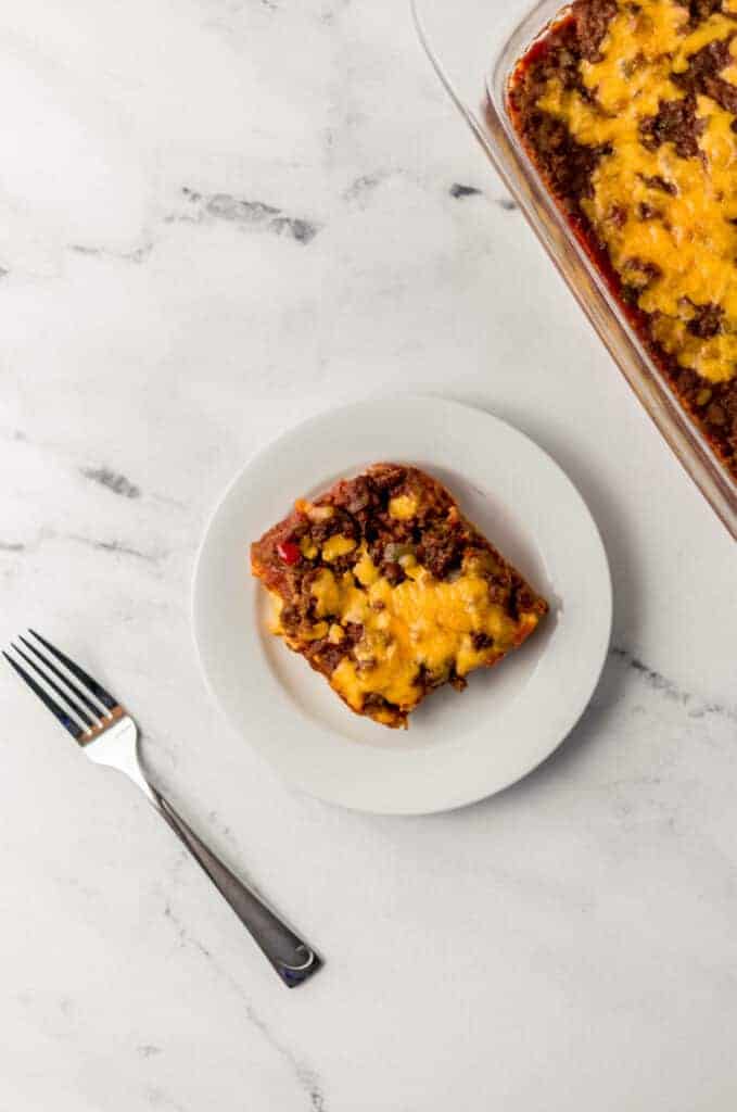 overhead view of sloppy joe cornbread casserole serving on a small white plate beside a fork and the rest of the casserole in a glass dish