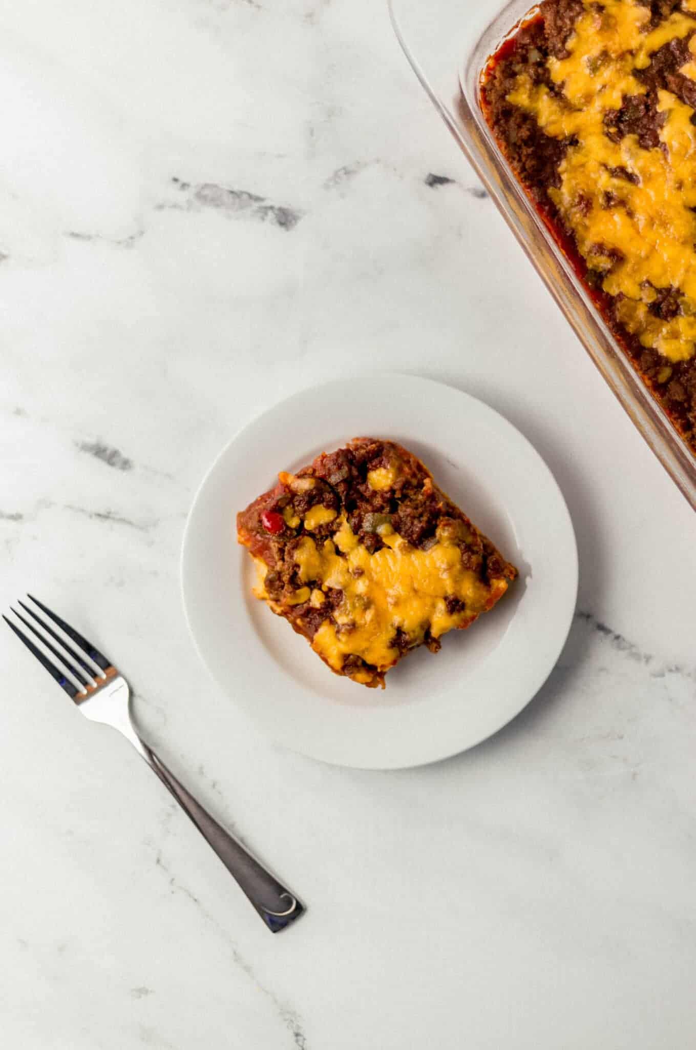 overhead view of sloppy joe cornbread casserole serving on a small white plate beside a fork and the rest of the casserole