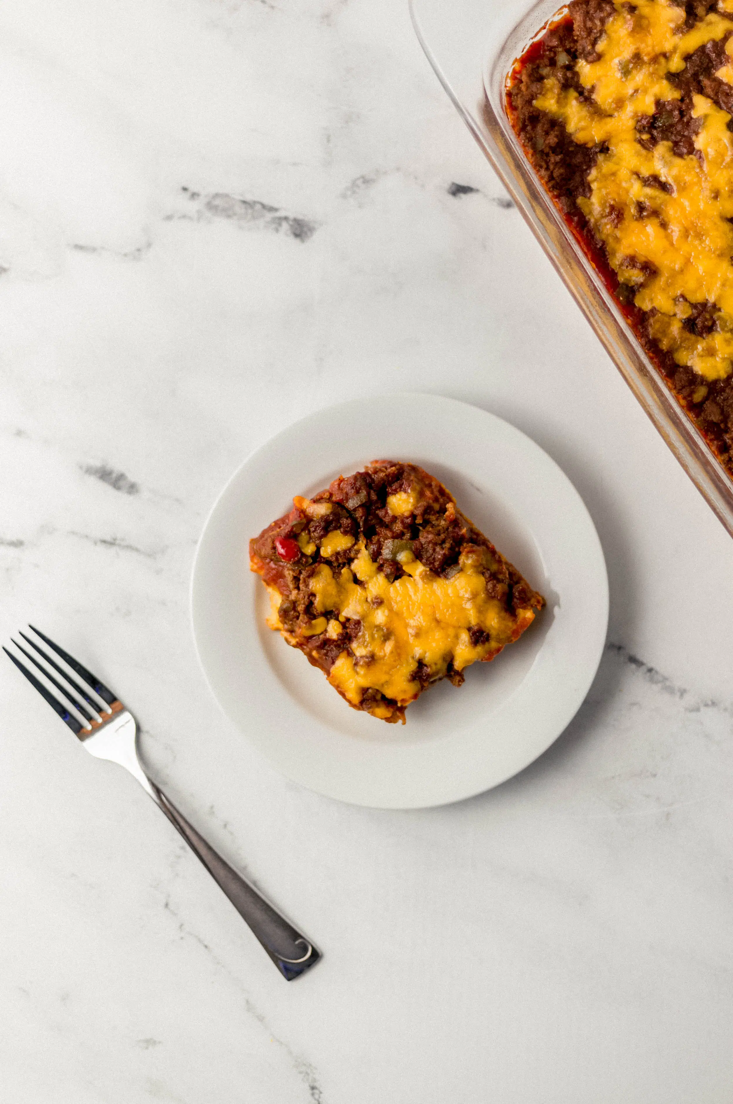 overhead view of sloppy joe cornbread casserole serving on a small white plate beside a fork and the rest of the casserole