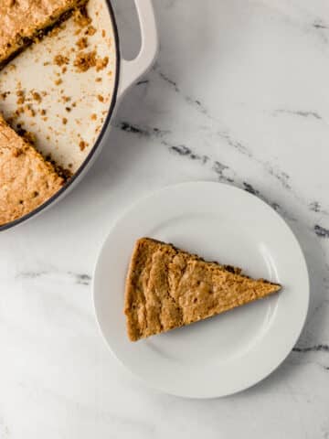 overhead view of piece of cookie on small white plate beside the rest of the cookie in the skillet