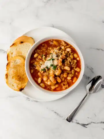 overhead view of white bowl of soup on white plate with bread beside a spoon