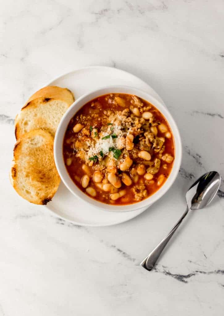 overhead view of white bowl of soup on white plate with bread beside a spoon