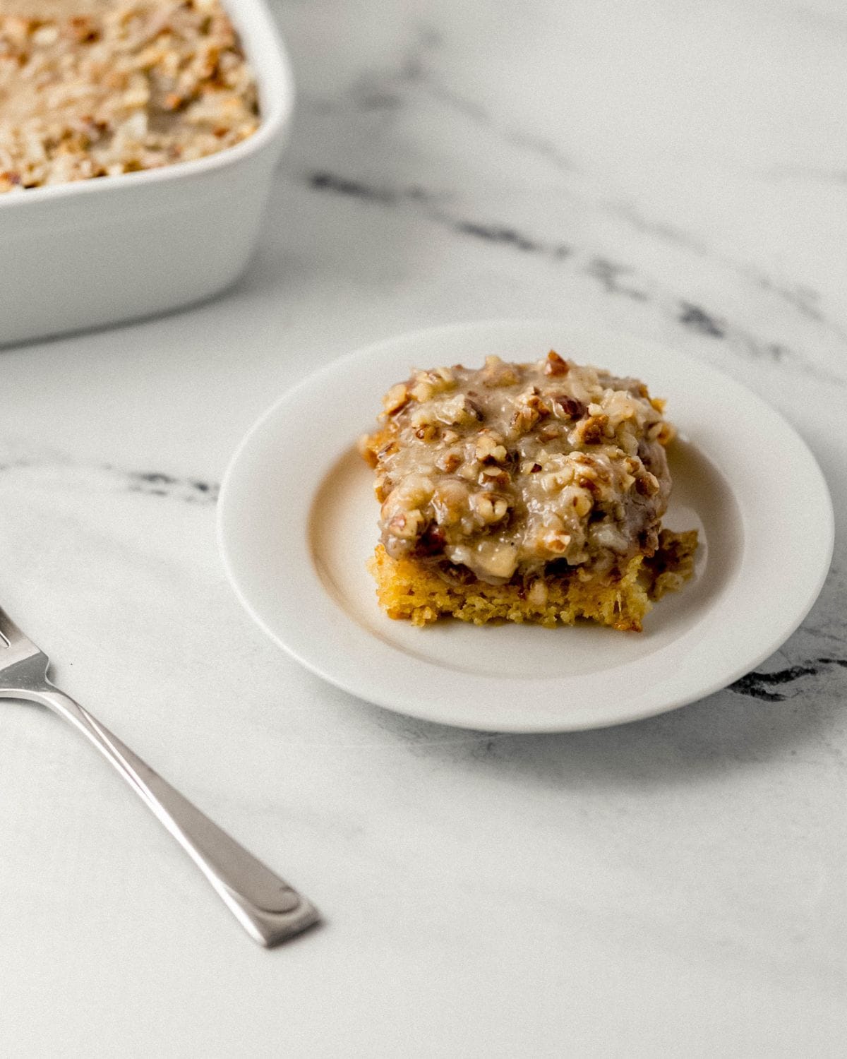 Side view of piece of cake on white plate by fork and the rest of the cake in the background. 