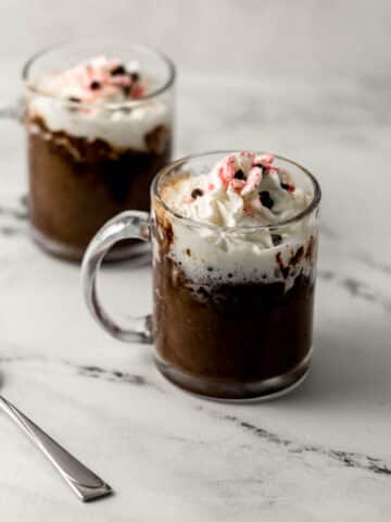 close up side view of two mug cakes topped with whipped cream, chocolate chips, and peppermint pieces