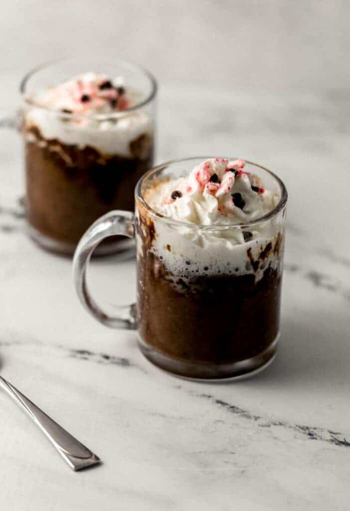 close up side view of two mug cakes topped with whipped cream, chocolate chips, and peppermint pieces