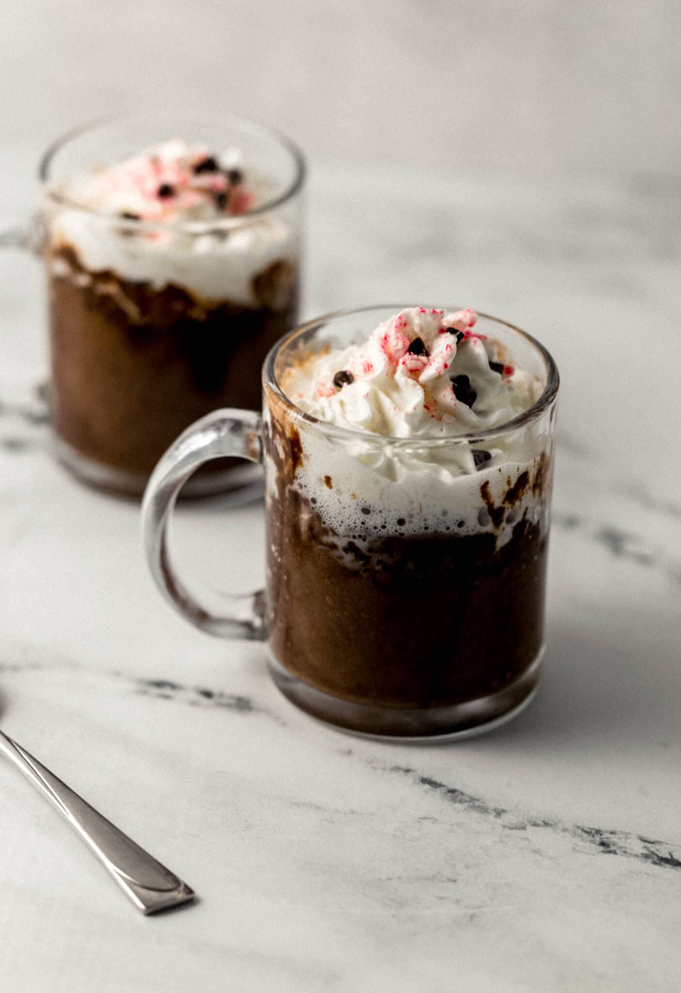 two finished mug cakes topped with whipped cream, peppermint, and chocolate chips 