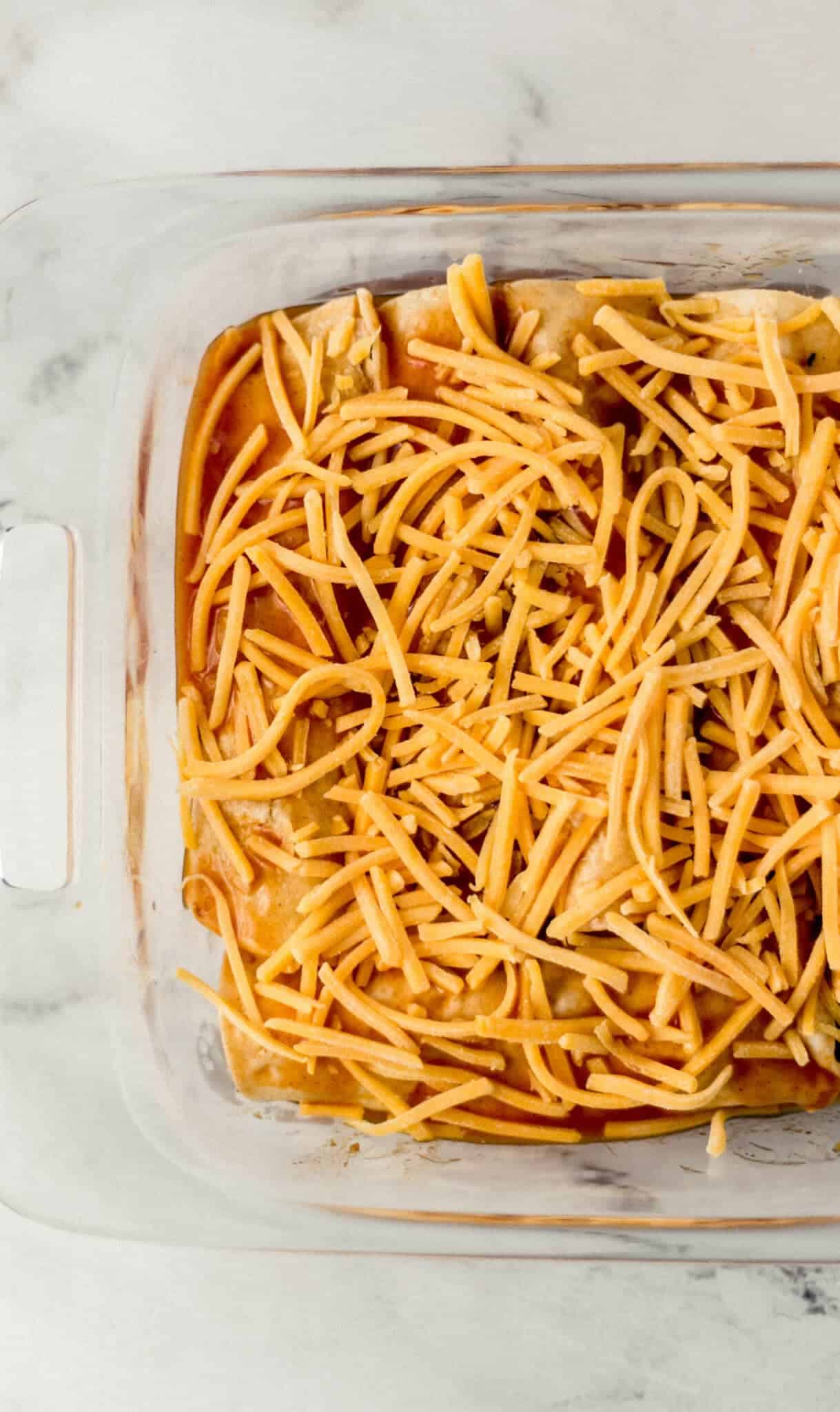 unbaked enchiladas topped with shredded cheese in glass baking dish 