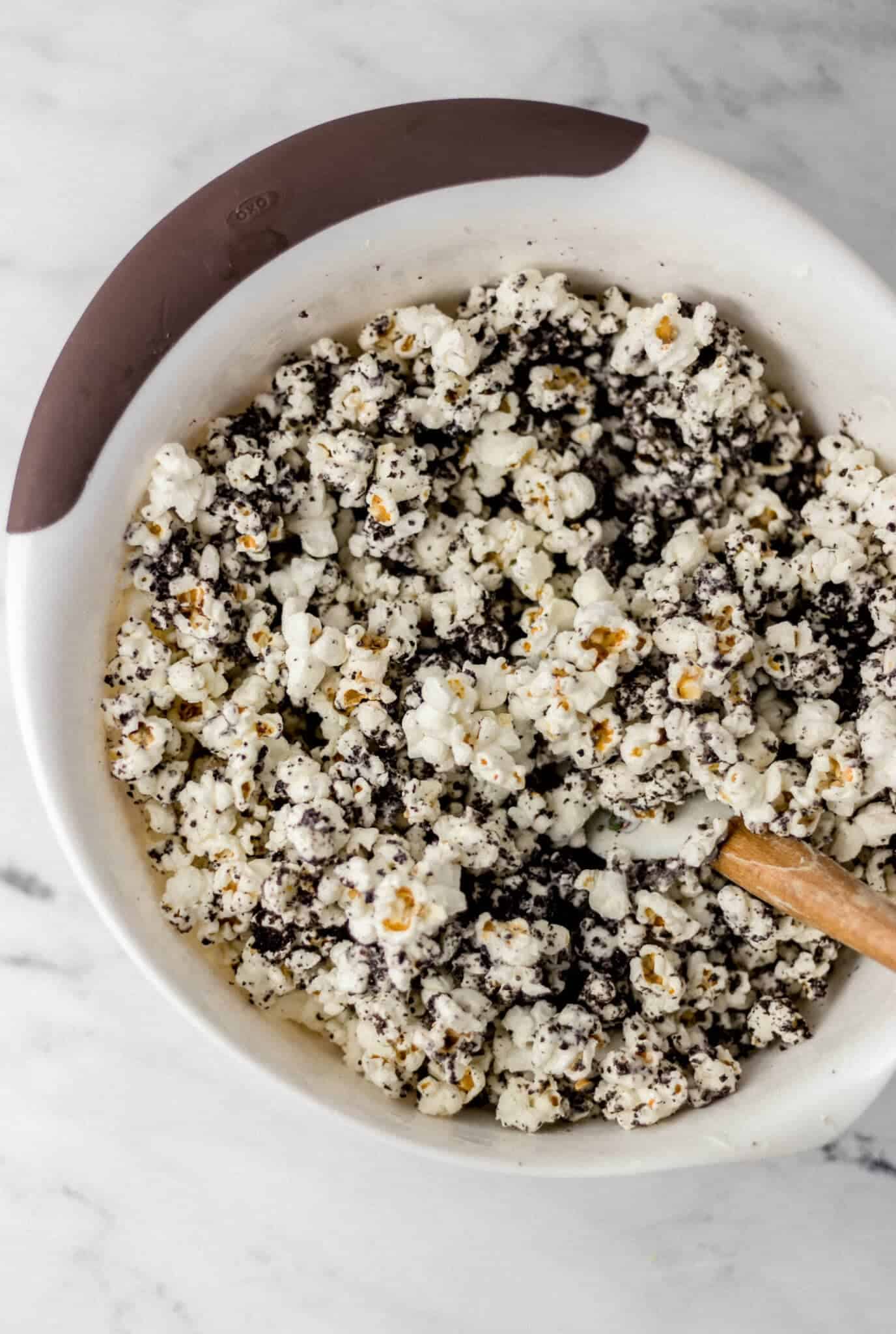 crushed cookies and melted chocolate added to popcorn in large white bowl with spatula