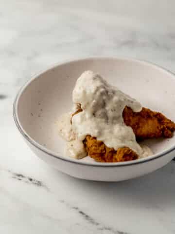 close up side view of chicken fingers topper with gravy in white bowl