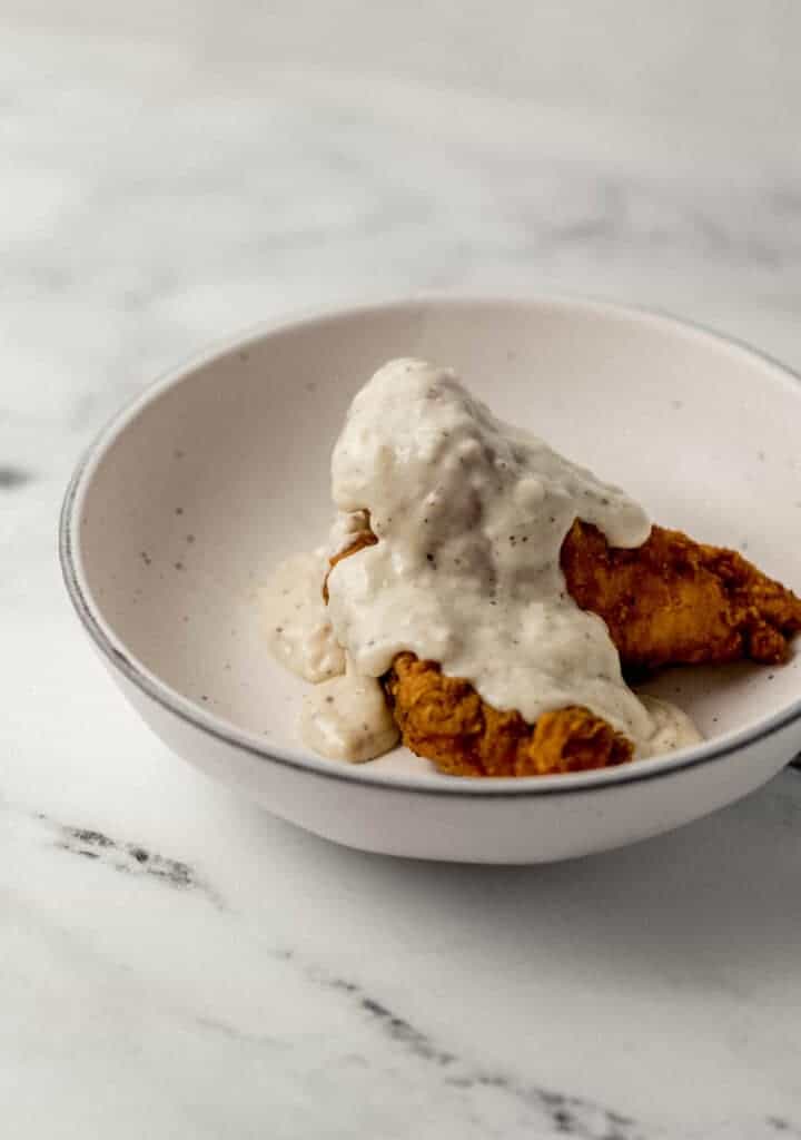 close up side view of chicken fingers topper with gravy in white bowl