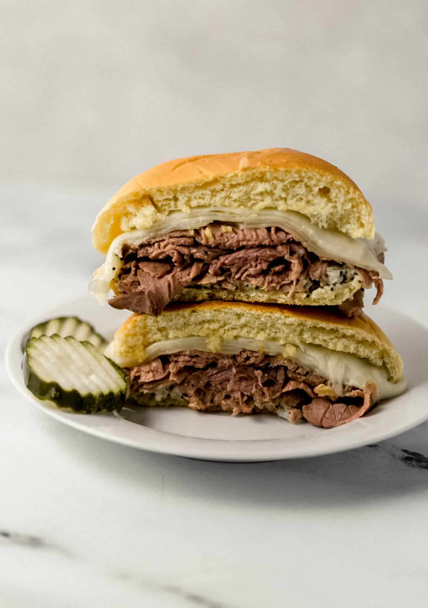 single roast beef sandwich on white plate, cut in half, and stacked on each other with pickles on the side 