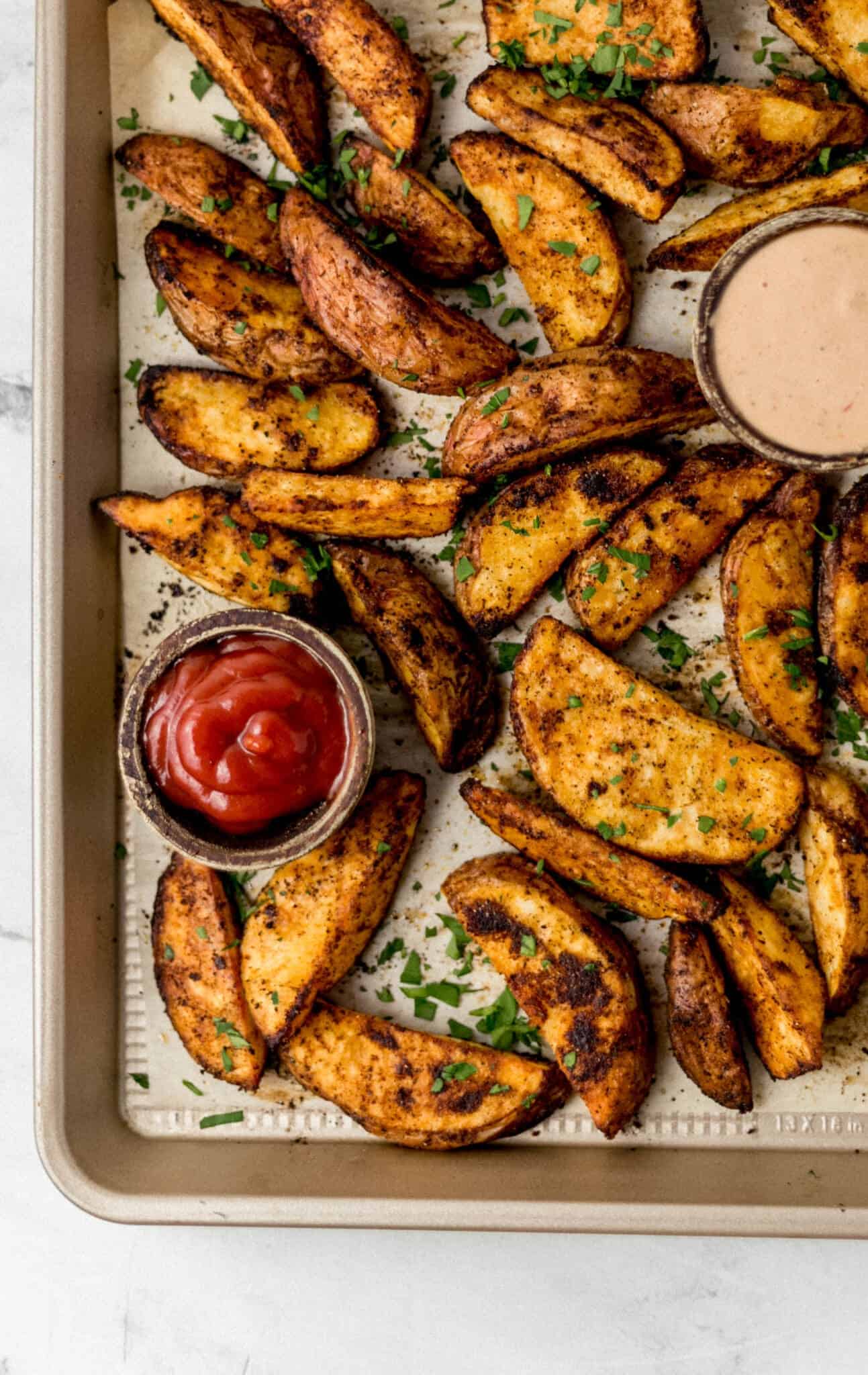 overhead view of finished potato wedges on parchment lined baking sheet with bowls of ketchup and sauce