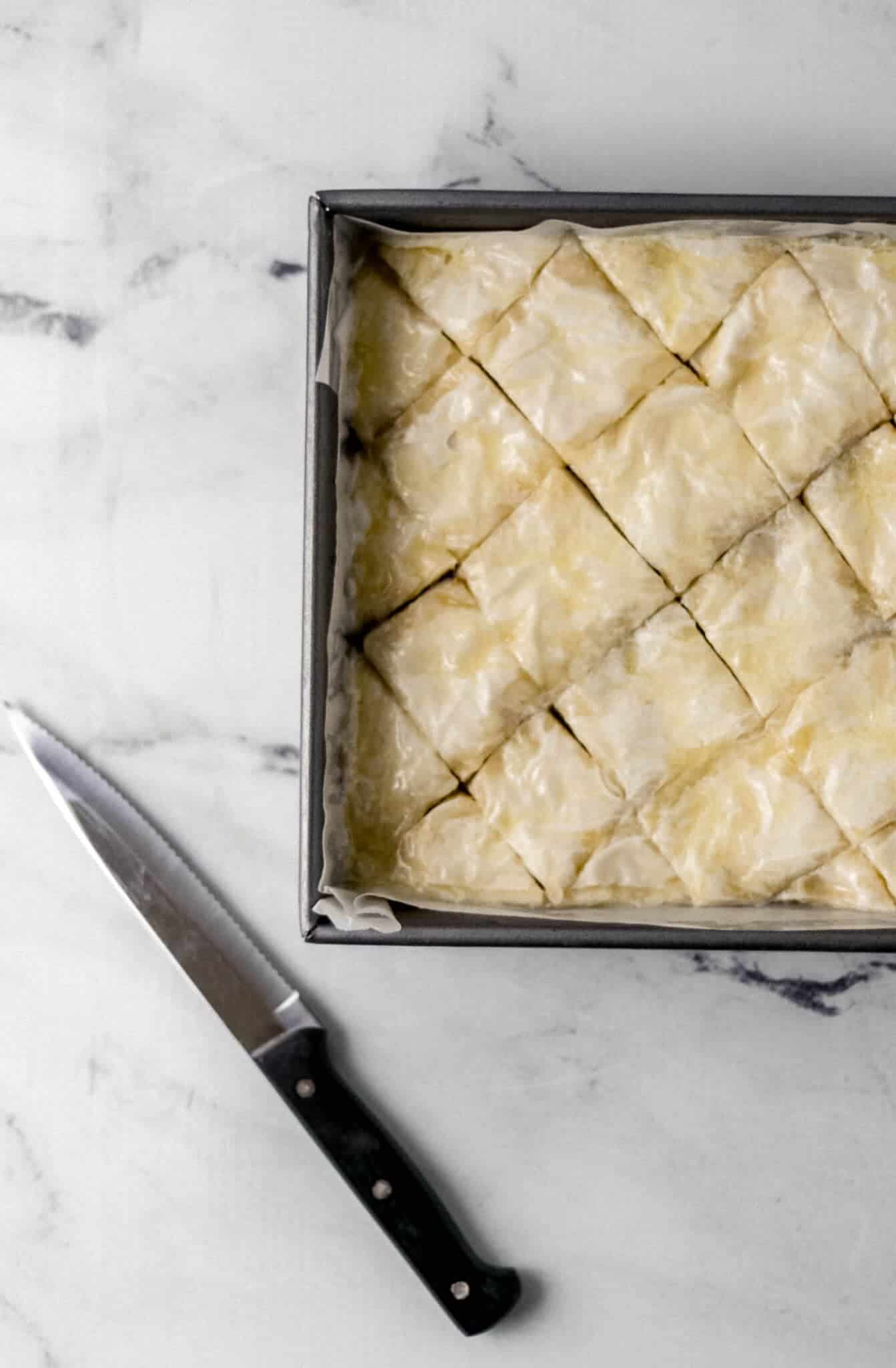 unbaked baklava cut into diamond shapes in pan beside a serrated knife on marble surface 